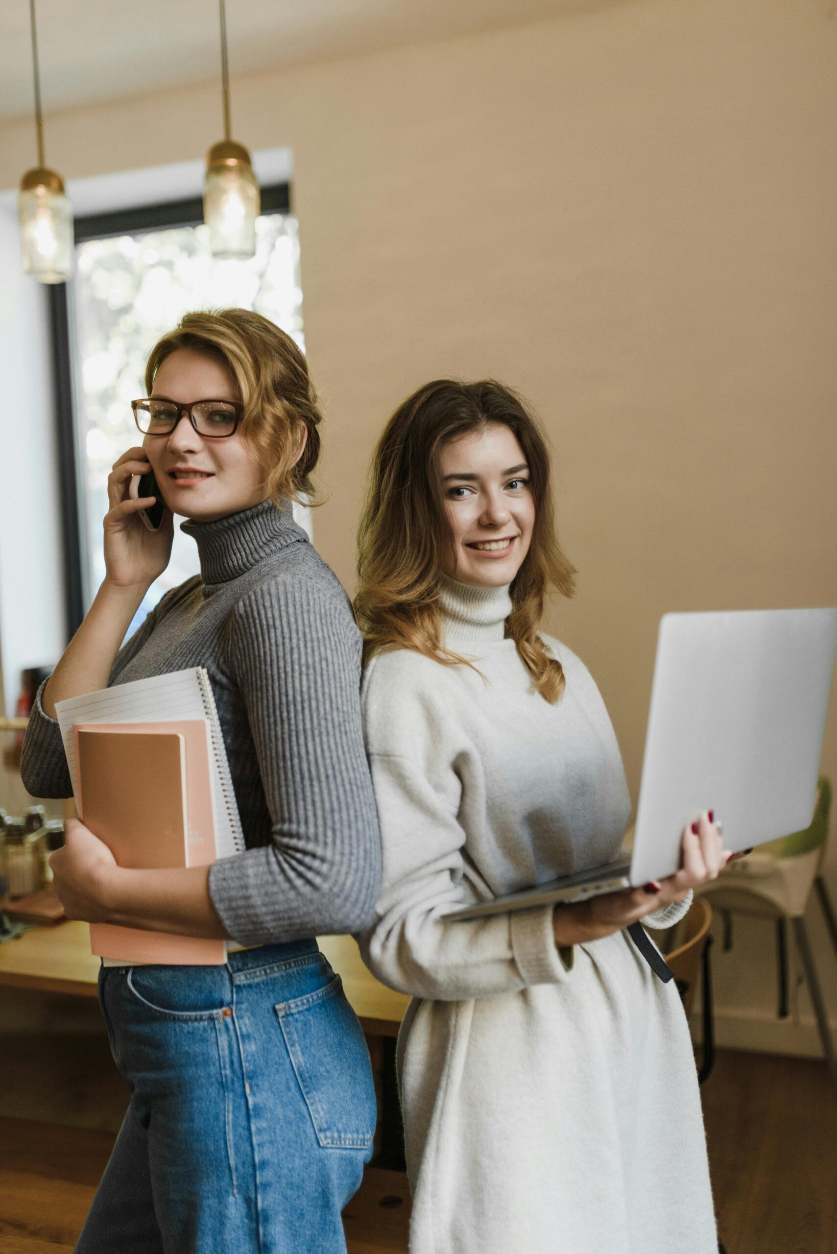 Two women stand back to back in an office, using laptops and mobile phones for work.